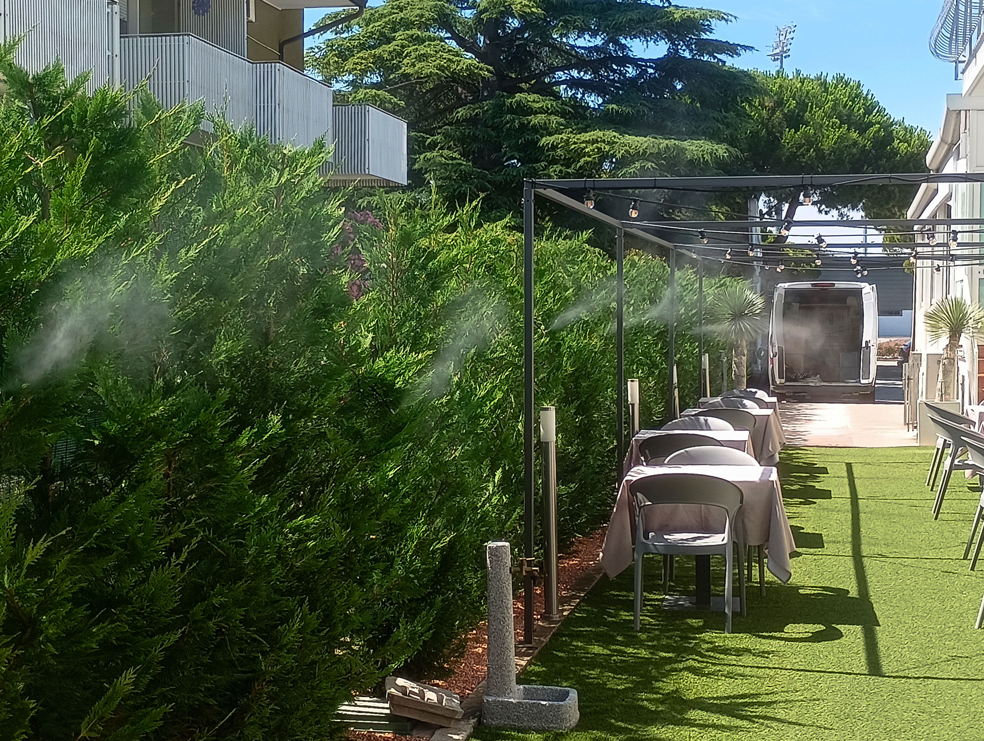 Sistema di nebulizzazione antizanzare installato lungo la siepe di un ristorante con tavoli all’aperto, visibile il getto fine di vapore tra le piante.