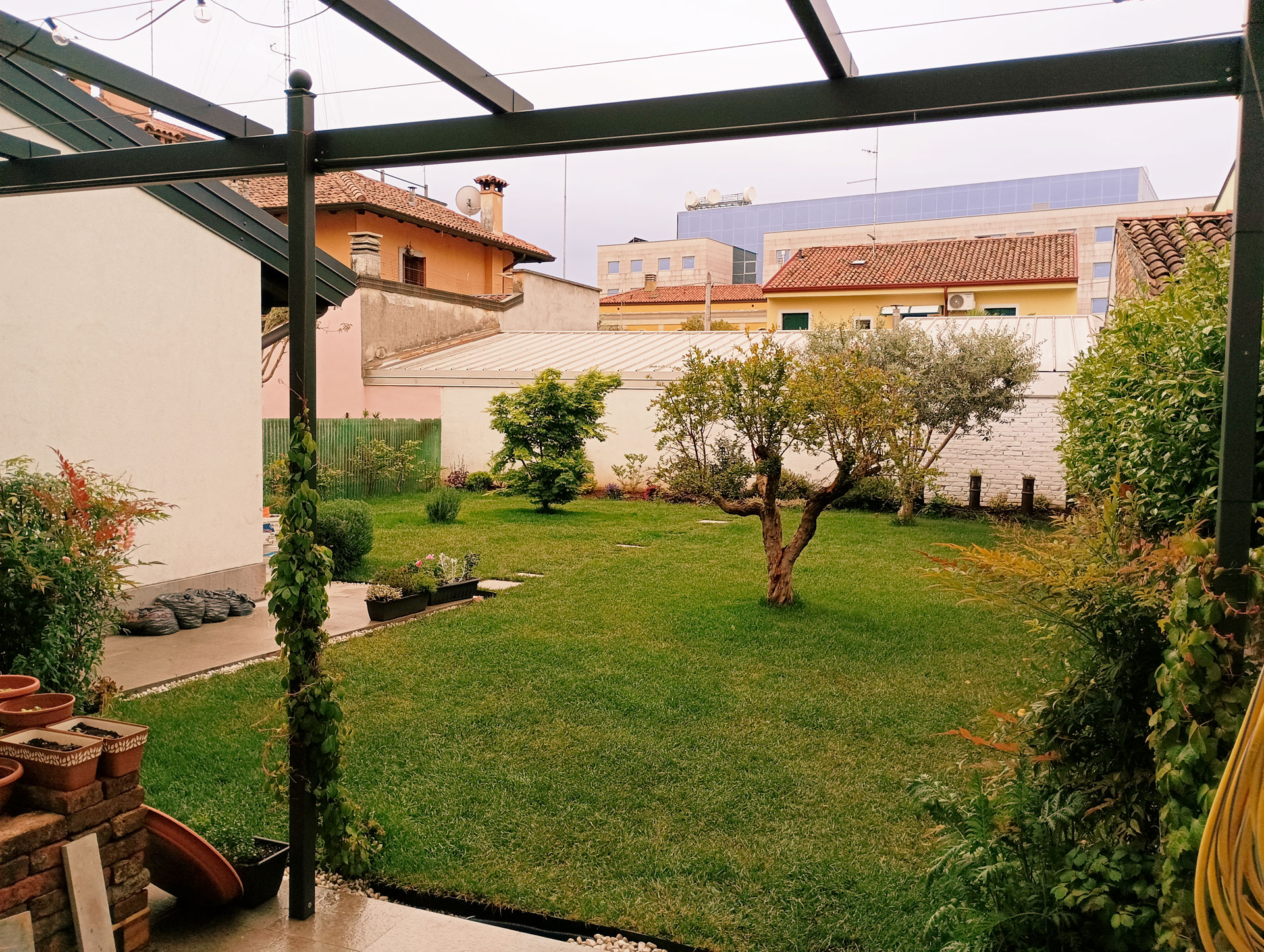 Giardino urbano privato con prato ben curato e alberi decorativi, visuale dalla veranda coperta di una casa con sistema antizanzare integrabile.